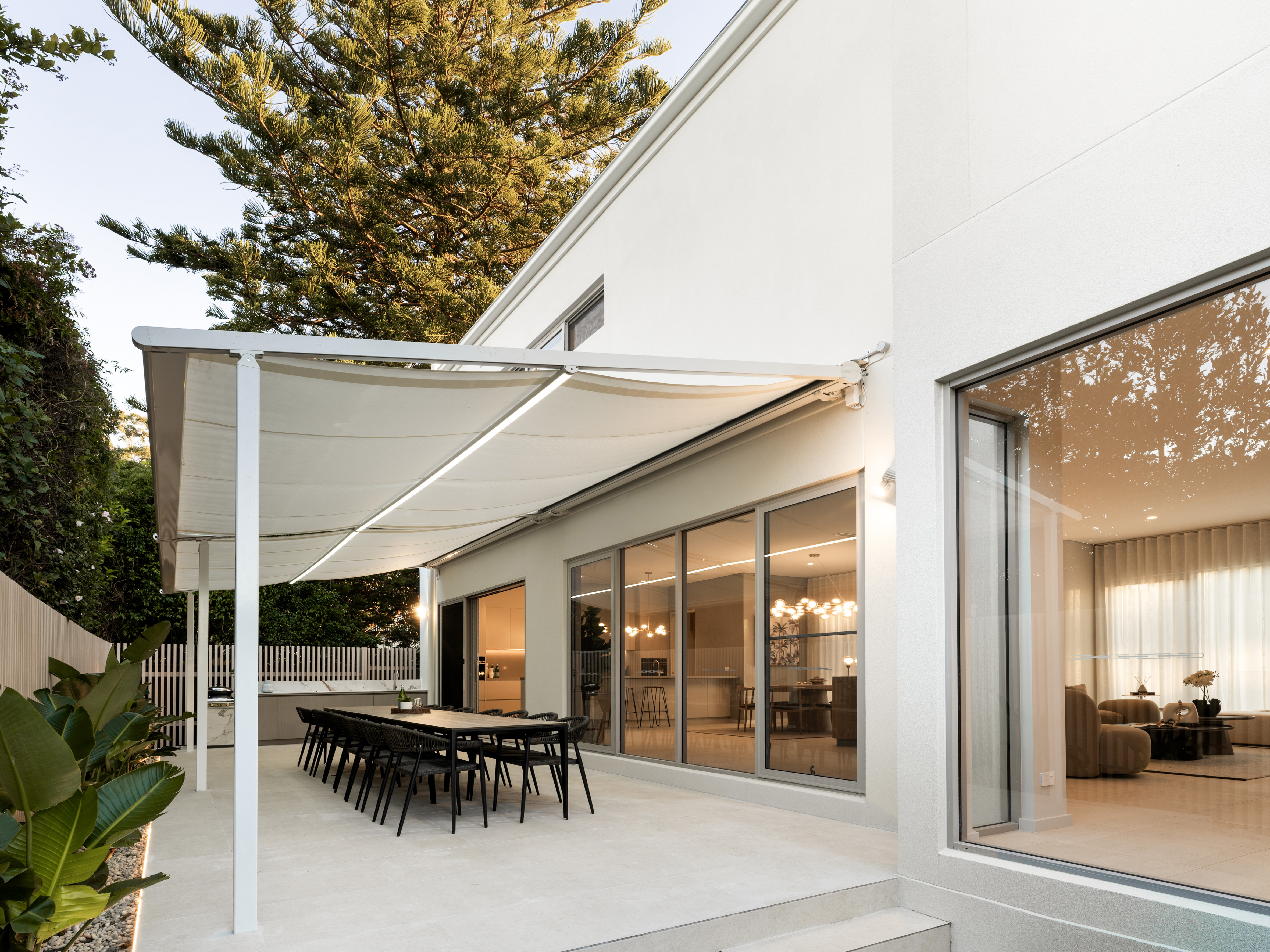 Modern outdoor patio with a long dining table, retractable canopy, and large windows looking into the home.